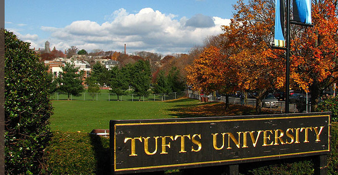 A large sign reading 'Tufts University' stands in front of a green lawn with autumn trees and campus buildings in the background under a partly cloudy sky.