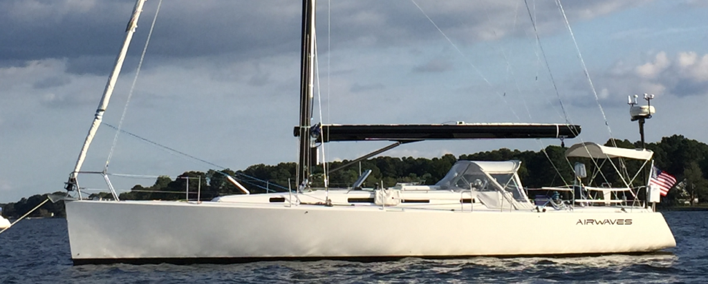 A white sailboat named 'AIRWAVES' floats on calm water near a tree-lined shore, with a partly cloudy sky above. An American flag is displayed at the stern.