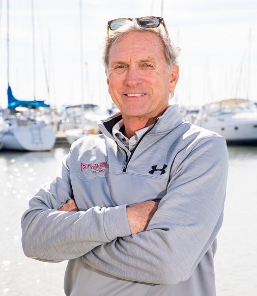 A man with gray hair stands in front of a marina with boats, wearing a gray quarter-zip jacket, sunglasses resting on his head, and smiling with his arms crossed.