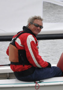 A person wearing a red and white jacket, sunglasses, and a life vest sits on a sailboat, smiling at the camera with water and a sail visible in the background.