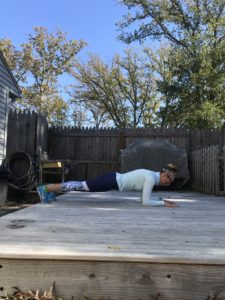 A person wearing athletic clothes and a cap holds a plank position on a wooden deck outside, with a grill and trees visible in the background on a sunny day.