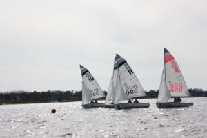 Three small sailboats with OCC markings race close together on a slightly wavy body of water; a red buoy floats nearby, and a shoreline with trees and houses is visible in the background under a cloudy sky.