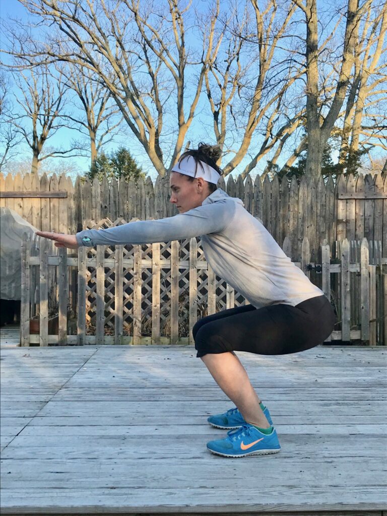 A woman in athletic clothing performs a squat exercise outdoors on a wooden deck, with bare trees and a wooden fence in the background.