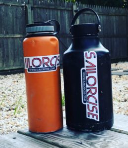 Two large water bottles, one orange and one black, both with "SAILORCISE" stickers, are placed on gravel outdoors near a wooden fence.