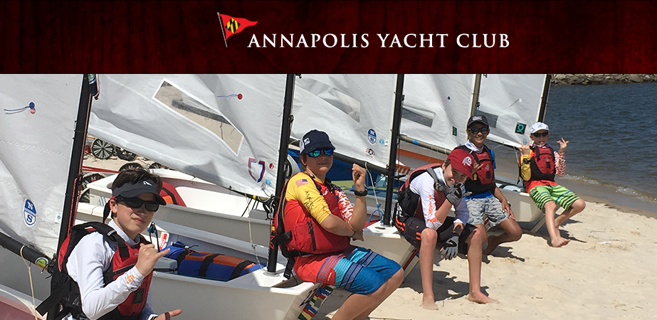 A group of kids wearing life jackets and sunglasses sit on small sailboats and sand at the beach, smiling and posing in front of the water. 'Annapolis Yacht Club' is written above them on a red banner.