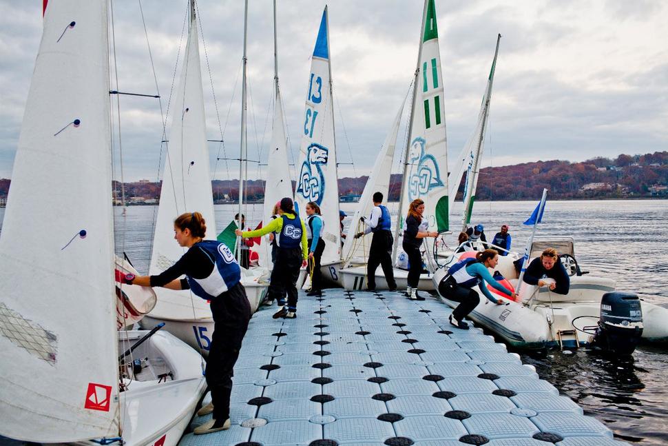 A group of people in sailing gear prepare sailboats on a floating dock, with calm water and a wooded shoreline in the background under a cloudy sky.