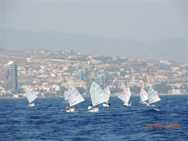 A group of small sailboats with white sails race on blue water near a coastal city, with hills and buildings visible in the background. The date “23 08 2018” is shown in the bottom right corner.