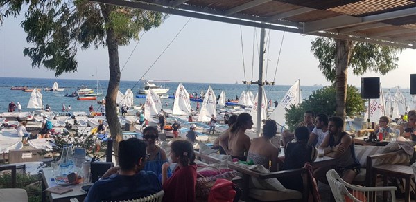 People sitting at a shaded outdoor café near the beach, with many small sailboats and people on the water in the background. The scene is lively and sunny, with trees providing partial shade.
