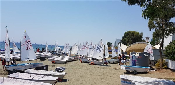 Small sailboats lined up on a sandy beach with sails raised, people preparing boats, and the sea visible in the background under a clear blue sky. Trees and buildings are on the right side of the image.
