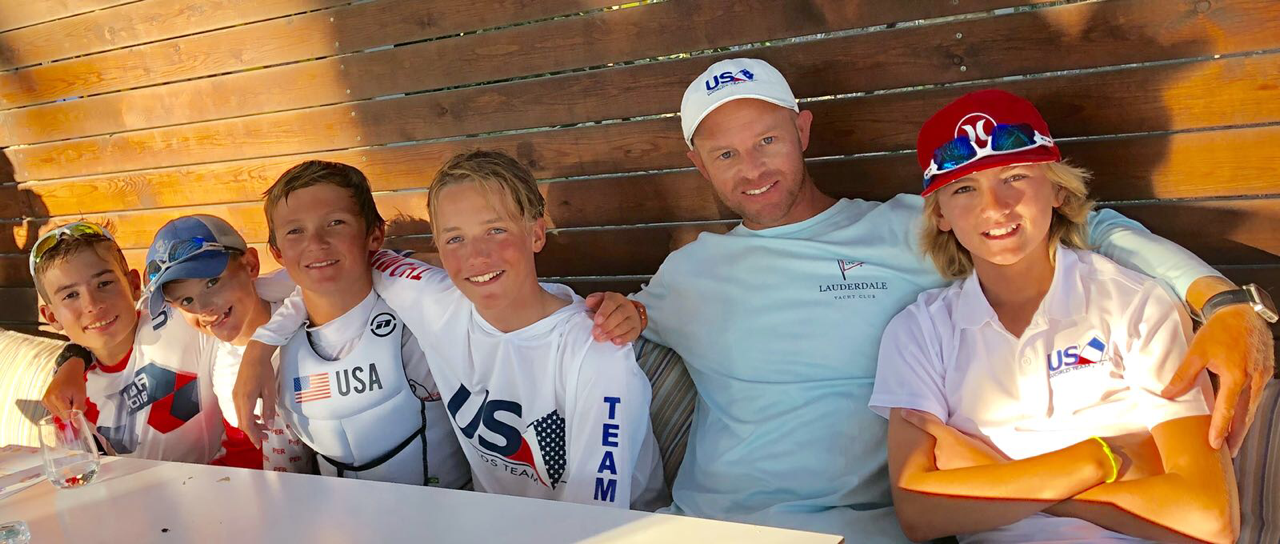 A group of five boys and one adult, all wearing sailing or USA-themed shirts and caps, sit closely together on a bench, smiling under warm sunlight against a wooden wall.