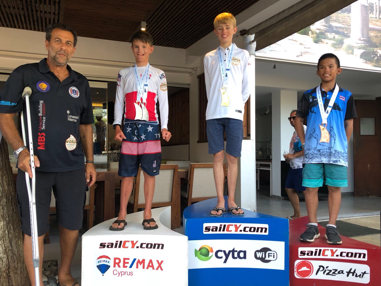 Four boys at a sailing competition award ceremony: one stands on crutches beside the podium, while the other three stand on first, second, and third place podiums, each wearing medals and smiling. Sponsor logos are on the podiums.