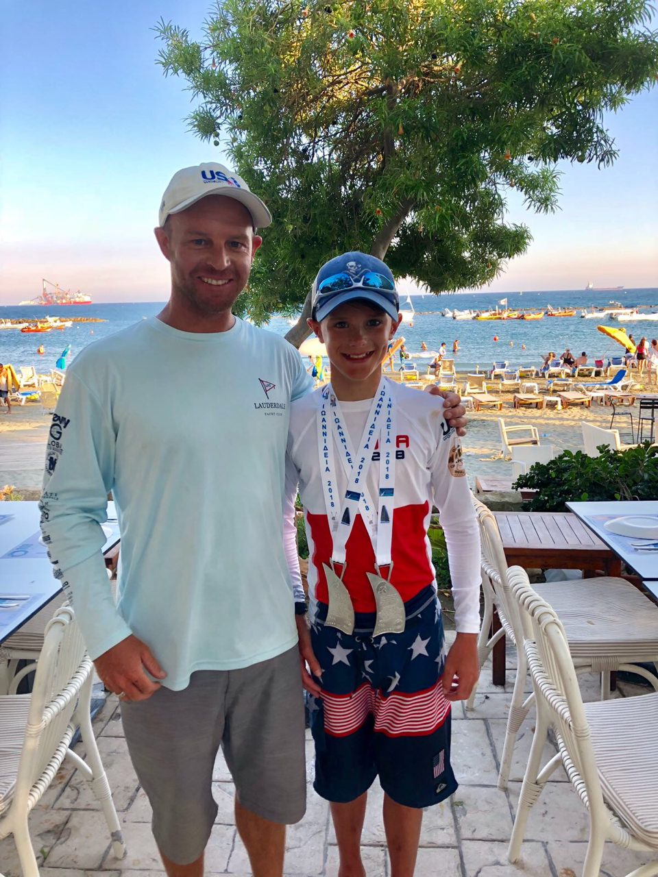 A smiling man and a boy stand together outside by the sea. The boy wears patriotic clothing, a cap, and two medals around his neck. Behind them are tables, chairs, and people enjoying a sunny beach.