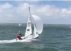 Two people sail a small white racing dinghy with the number 805 on the sail, speeding across choppy blue water under a partly cloudy sky.