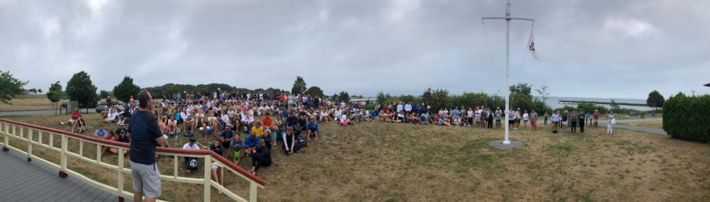 A large crowd sits on a grassy lawn near a flagpole, listening to a speaker on a porch. Trees and water are visible in the background under a cloudy sky.
