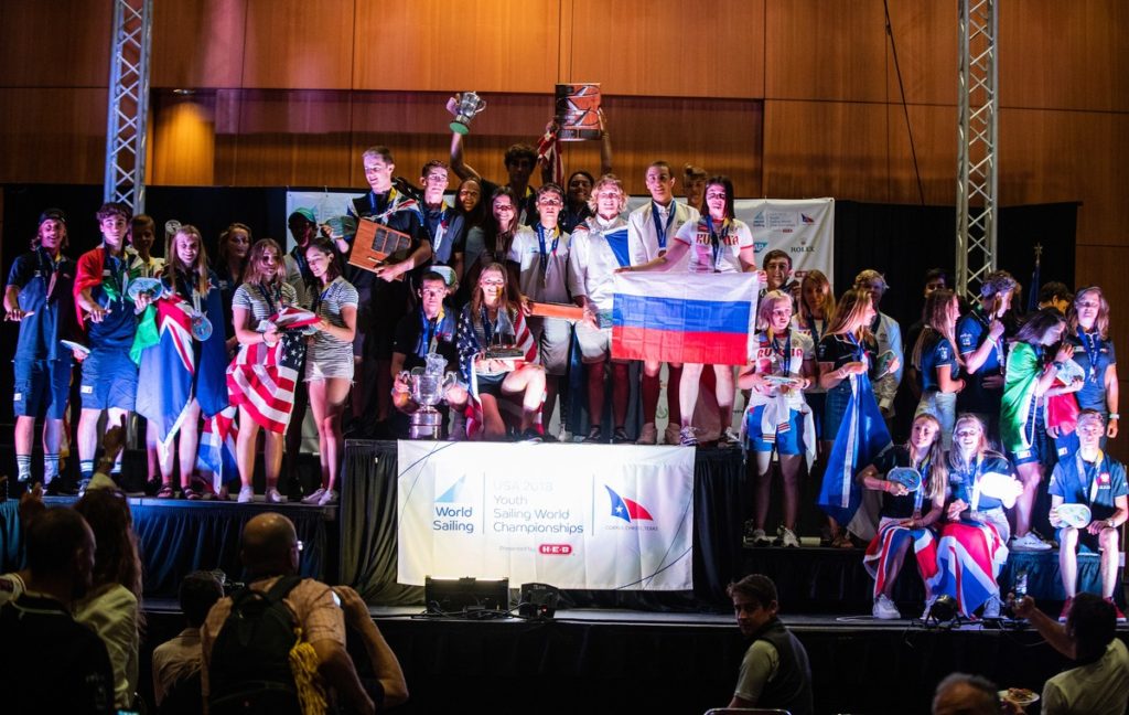 A large group of young athletes stands on a podium at a sailing championship award ceremony, holding trophies and flags from different countries, with a crowd watching in front.