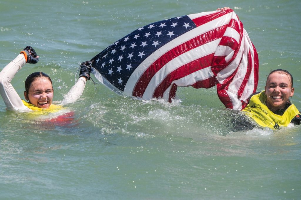 Two smiling people in yellow life vests swim in the water, holding up a large American flag above the surface.