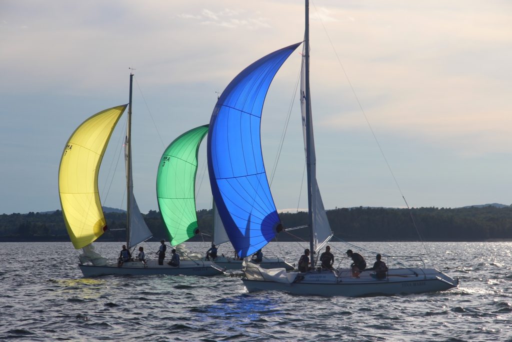 Three sailboats race side by side on a calm body of water, each with a colorful spinnaker sail—yellow, green, and blue—against a backdrop of distant hills and a partly cloudy sky.
