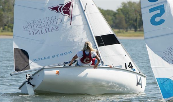 A person with long blonde hair, wearing a life jacket, sails a small white sailboat marked with the number 14. The boat features “College of Charleston Sailing” on the sail, and another sailboat is partially visible nearby.