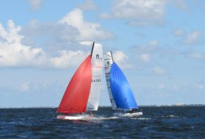 Two sailboats race on open water under a partly cloudy sky; one has a red sail and the other has a blue sail, with land visible in the distant background.