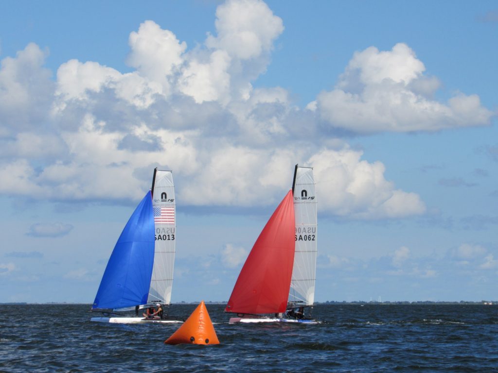 Two small sailboats race on the water, one with a blue sail and the other with a red sail, passing an orange triangular buoy under a bright, partly cloudy sky.