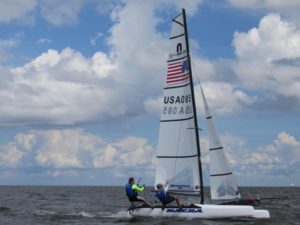 Two people sailing a small catamaran with the sail marked "USA 085" on a body of water under a partly cloudy sky. Both are wearing life jackets and leaning out to balance the boat.