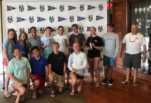 A group of teens and an adult stand indoors, posing for a photo in front of a backdrop with SYC and S.S. logos. Some hold drink tumblers, and everyone is casually dressed, smiling at the camera.