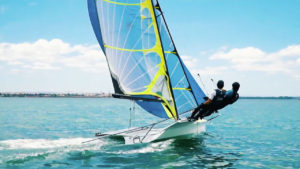 Two people sail a small boat with colorful blue and yellow sails on a calm, clear blue sea under a bright sky. They are leaning out over the water to balance the boat as it speeds forward.