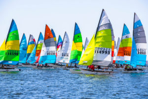 Colorful sailboats with multi-colored, patterned sails race closely together on blue water under a clear sky, creating a vibrant and energetic scene. Sail numbers and crew members are visible on many of the boats.