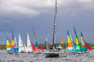 A group of colorful sailboats gathers at the start line near a larger boat with flags and people on board, under a cloudy sky on a body of water.
