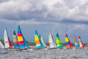 A group of colorful sailboats with vibrant sails participate in a race on a lake under a cloudy sky, with green trees visible in the background.
