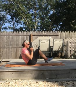 A person in athletic wear performs a seated yoga pose on an orange mat outdoors, with hands clasped, knees bent, and feet off the ground. Wooden fencing and trees are in the background.