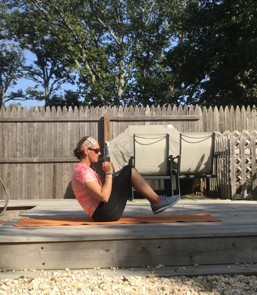A person in athletic wear performs a seated yoga pose on an orange mat outdoors, with hands clasped, knees bent, and feet off the ground. Wooden fencing and trees are in the background.