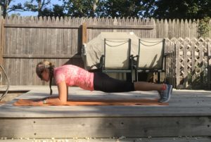 A person wearing a pink shirt, black leggings, and gray sneakers is doing a plank exercise on an orange mat outdoors on a wooden deck, with a fence and some outdoor furniture in the background.