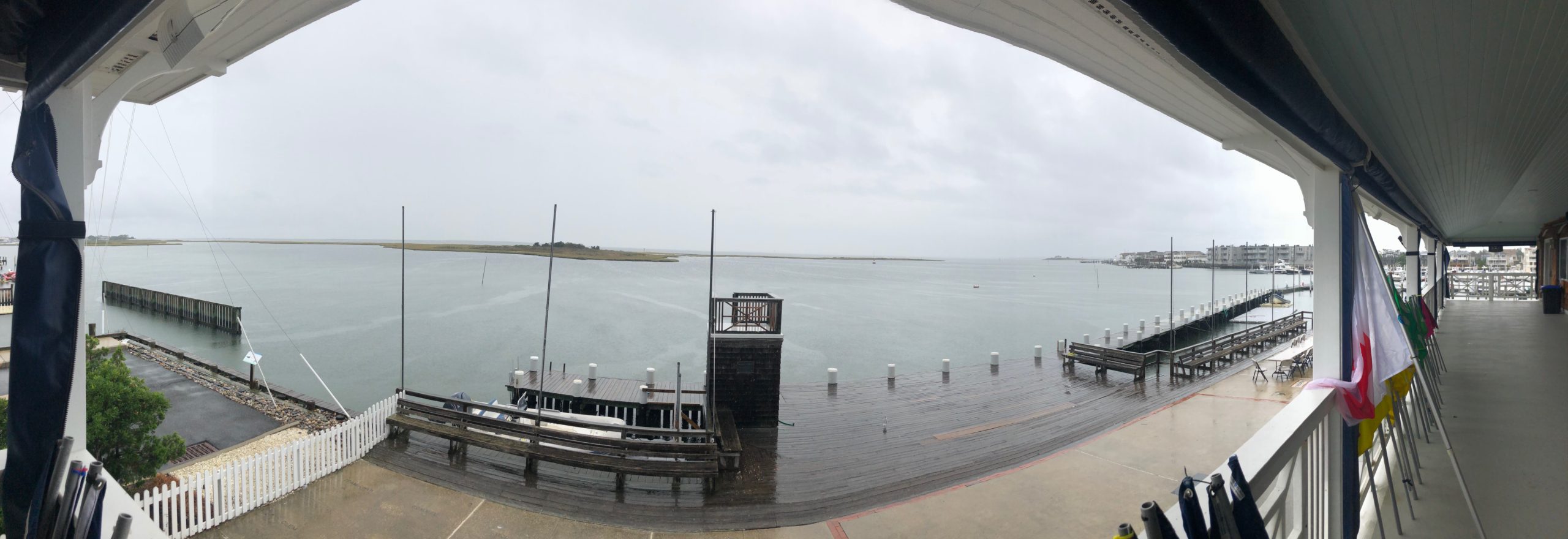 Panoramic view from a covered porch overlooking a calm bay with wooden docks, benches, and a white fence on a cloudy, gray day. Buildings and boats are visible in the distance across the water.