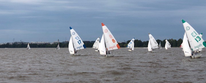 Several small sailboats with colorful numbered sails race across a wide brown river under a cloudy sky, with green trees and distant buildings visible on the far shore.