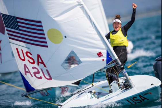 A sailor wearing a yellow vest and headband celebrates on a small sailboat with 'USA' and an American flag on its sail, during a sunny day on the water.