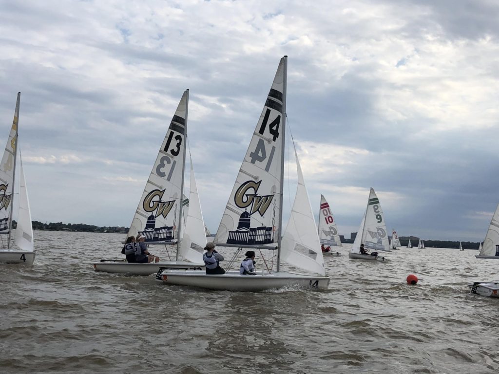 Several small sailboats with white sails and numbers, including "13" and "14," race on a cloudy day. Crews in life vests steer on choppy water, with other boats and an orange buoy visible in the background.