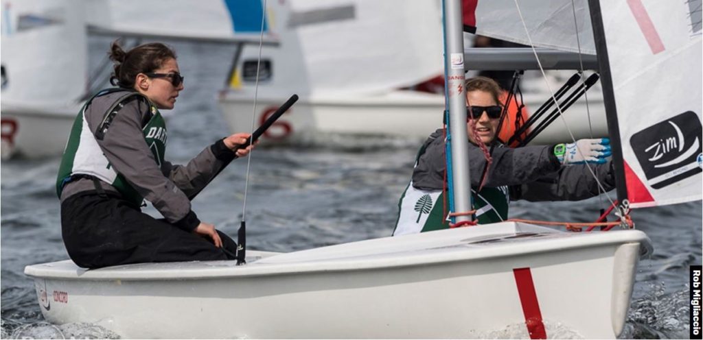 Two sailors wearing sunglasses and life jackets compete in a small sailboat race on the water, focusing intently as they steer and adjust the sails. Other boats and competitors are visible in the background.