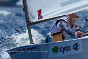 A young sailor wearing sunglasses and gear steers a small sailboat through choppy blue water during a sailing competition. The sailboat displays sponsorship logos and the sailor's focused expression is visible.