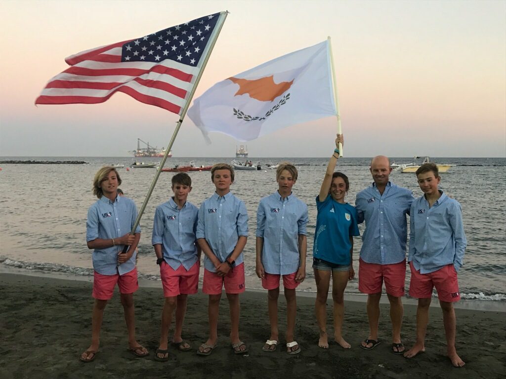 Seven people stand on a beach at sunset, wearing blue shirts and pink shorts. Two hold large flags: one of the United States, the other of Cyprus. Boats are visible in the water in the background.