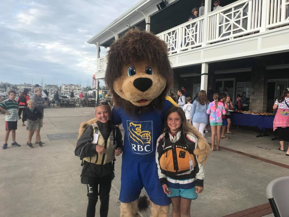 A person in a lion mascot costume with an "RBC" shirt stands outside with two smiling girls in life jackets on either side, among a crowd near a building and marina.