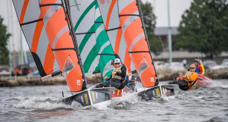 Three people sailing small boats with orange and green sails on choppy water, competing in a regatta near a shoreline with trees and parked cars in the background.