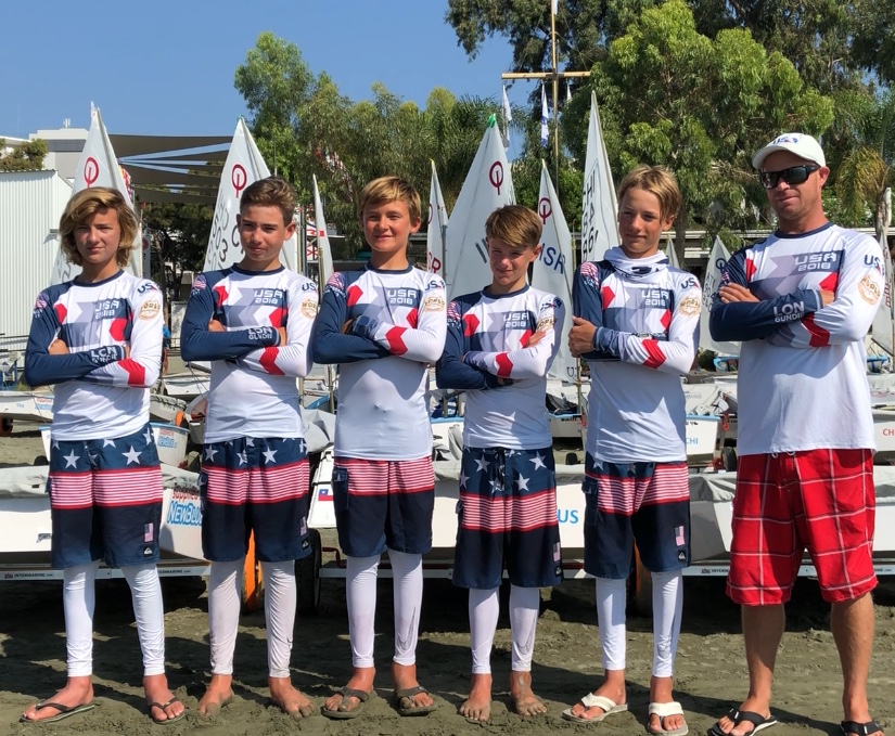 Six people, five youth and one adult, stand side by side in matching USA sailing uniforms in front of small sailboats. They are outdoors, with trees and boats visible in the background.