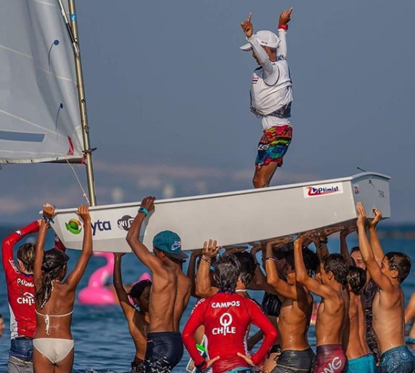 A group of children in swimsuits and red shirts hold a small sailboat above their heads in the water while a child stands on the boat, raising both middle fingers in the air.