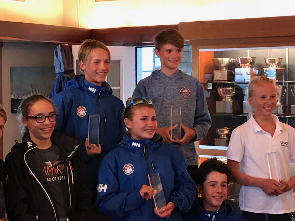 Six smiling teenagers, some wearing blue sailing jackets and holding clear rectangular trophies, pose indoors in front of a trophy display case. One wears a white polo, and another wears glasses and a black jacket.