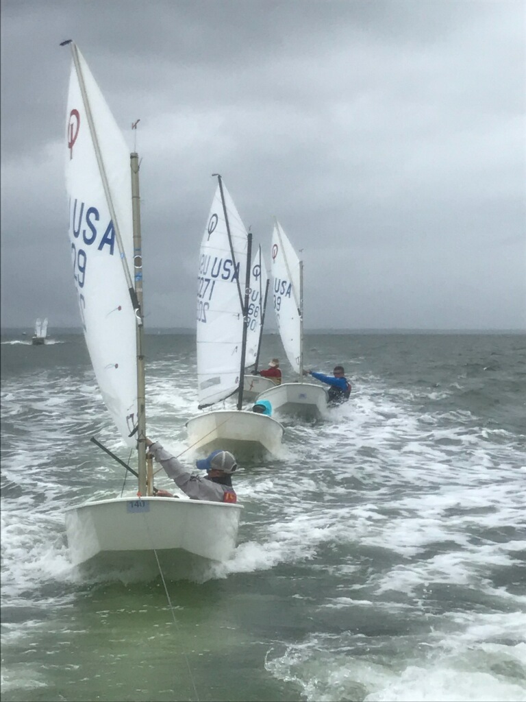 Three small sailboats with white sails, each carrying one person, sail closely together on choppy gray water under a cloudy sky. The sailboats have “USA” and numbers printed on the sails.