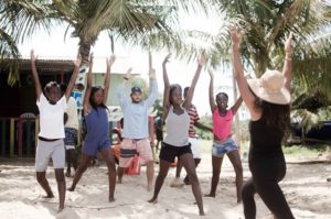 A group of people, including children and adults, stand on a sandy beach with their arms raised, following an instructor in a sunhat. Palm trees and colorful buildings are in the background.