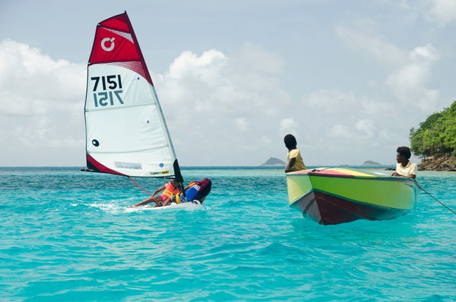 Two children in a colorful boat watch another child sailing a small red and white sailboat on bright turquoise water near a lush, green island under a partly cloudy sky.