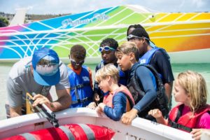A group of children wearing life jackets and swim goggles gather around an adult demonstrating something in a boat, with a colorful boat labeled “Precious Fire” in the background.