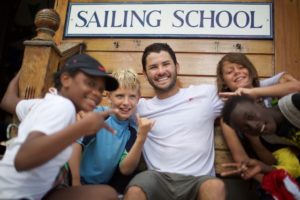 A smiling man sits on wooden steps under a "Sailing School" sign, surrounded by five happy children making playful hand gestures, all appearing cheerful and excited.